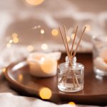 Home perfume in glass bottle with wood sticks, scented burn candles  tray in bedroom close up. Aromatherapy cozy atmosphere lifestyle. Winter warm xmas season.